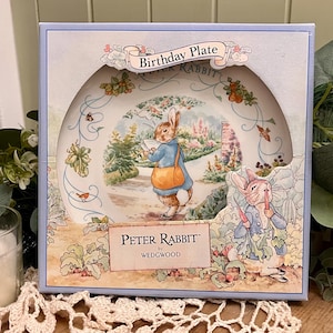 May include: A Peter Rabbit birthday plate in a decorative box. The plate features an illustration of Peter Rabbit in a blue jacket and orange satchel. The box has the words "Birthday Plate" and "Peter Rabbit" printed on it.