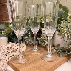 May include: Three clear champagne flutes with diamond-cut patterns, displayed on a wooden board. The flutes have a classic design with a wide bowl and a long stem. The background includes greenery and a candle.