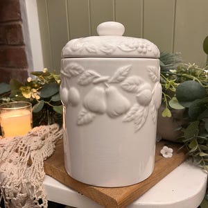 May include: A white ceramic storage jar with a lid, decorated with embossed fruit and leaf motifs. The jar is cylindrical and rests on a wooden board. The lid has a small handle and a floral design. The style is rustic and decorative.