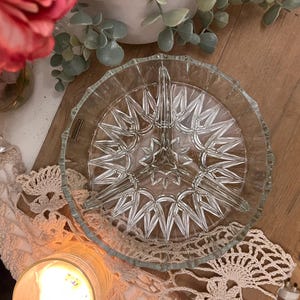 May include: A clear glass serving dish with a starburst design. The dish has three sections and a scalloped edge. The dish is on a wooden surface, with a candle and flowers in the background.