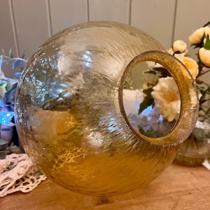 May include: A spherical, textured glass vase with a large circular opening. The vase has a golden hue and a reflective surface. The opening is framed with a gold-coloured rim. Decorative flowers are visible in the background.