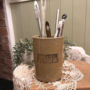 May include: A beige ceramic kitchen utensil holder with the words "KITCHEN UTENSILS" printed in black. It contains various stainless steel kitchen tools. The holder sits on a wooden surface covered with a white lace doily.