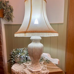 May include: A vintage table lamp with a cream lampshade and a decorative fringe. The lamp base is a speckled beige colour. The lamp is on a small wooden table with a lace doily.