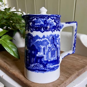 May include: A blue and white ceramic jug with a lid. The jug features a detailed landscape design. The handle is white with blue accents. The jug is sitting on a wooden tray.