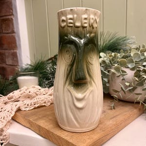 May include: A ceramic vase with a celery stalk design. The vase is off-white with green accents and the word "CELERY" is embossed at the top. The vase has a face-like design in the center. The vase is on a wooden surface.