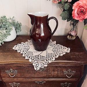 May include: A dark brown ceramic jug with a white interior, set on a white lace doily. The jug has a handle and a spout. It sits on a wooden chest of drawers with decorative handles. A vase of pink roses is in the background.