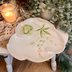 May include: A white, three-sectioned serving dish with a floral design. The dish has a green ceramic frog figurine and is decorated with green leaves and pink flowers. The dish is on a small white table with a candle and greenery in the background.