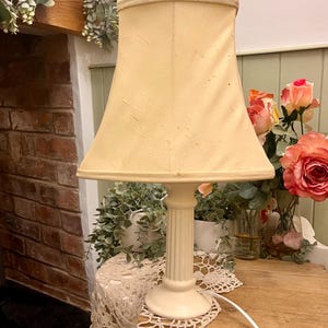 May include: Cream-coloured table lamp with a fabric lampshade. The lamp has a fluted column base and a round foot. The lampshade is a light beige colour. The lamp is on a small wooden table with a lace doily.