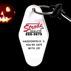 May include: A white key tag with the text "Strode Realty 555-3679 Haddonfield, IL YOU'RE SAFE WITH US!" and a set of keys. A carved pumpkin with a glowing face is in the background.