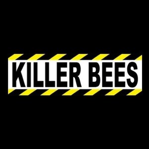 May include: A rectangular sign with the words "KILLER BEES" in bold, black capital letters. The sign has a white background with yellow and black diagonal stripes along the top and bottom edges, set against a black background.