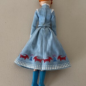 May include: A doll wearing a light blue dress with a flared skirt and long sleeves. The skirt has a red and blue pattern along the hem. The doll has red hair and is wearing blue boots.