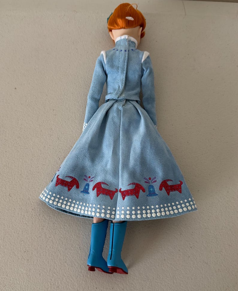 May include: A doll wearing a light blue dress with a flared skirt and long sleeves. The skirt has a red and blue pattern along the hem. The doll has red hair and is wearing blue boots.
