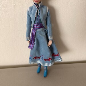 May include: A doll wearing a light blue dress with a matching jacket. The dress has a red and white pattern at the hem. The jacket has white trim and blue buttons. The doll is wearing blue boots and a purple sash.