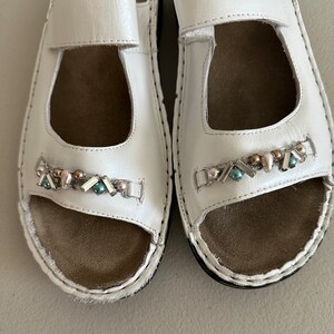 May include: A pair of white leather sandals with a decorative beaded strap. The sandals have a closed toe and an open heel.