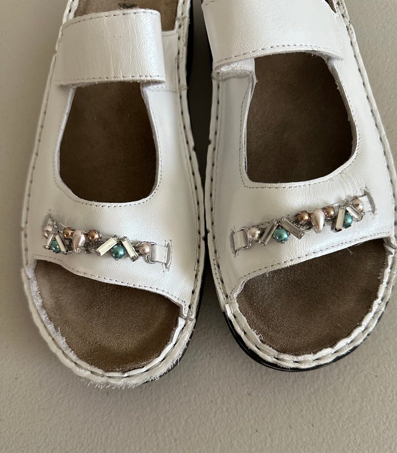 May include: A pair of white leather sandals with a decorative beaded strap. The sandals have a closed toe and an open heel.