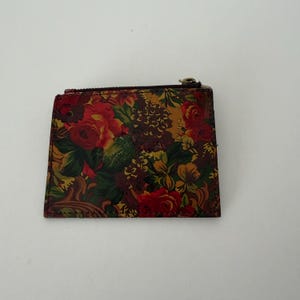 May include: A floral print wallet with a zipper closure. The wallet features a colorful pattern of red roses, green leaves, and other flowers on a dark background. The zipper is gold-toned.