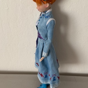 May include: A doll with red hair styled in an updo, wearing a light blue dress with white trim and decorative patterns. The doll's dress has a matching blue and purple sash. The doll is wearing blue boots and a decorative headpiece.