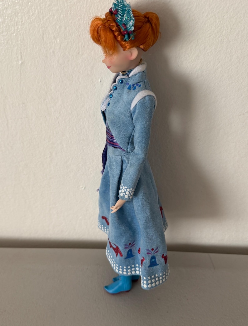 May include: A doll with red hair styled in an updo, wearing a light blue dress with white trim and decorative patterns. The doll's dress has a matching blue and purple sash. The doll is wearing blue boots and a decorative headpiece.