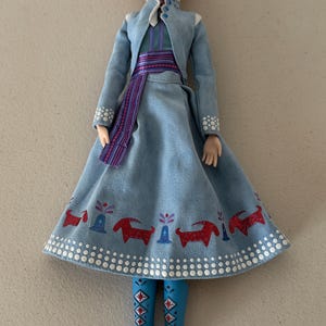 May include: A doll of a character with red hair, wearing a light blue dress and jacket. The dress has a pattern of red animals and blue boots. The doll has a blue and green hair accessory.