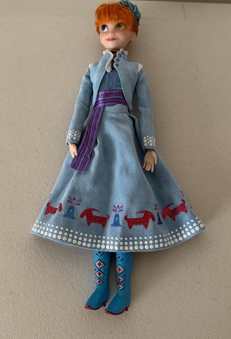 May include: A doll of a character with red hair, wearing a light blue dress and jacket. The dress has a pattern of red animals and blue boots. The doll has a blue and green hair accessory.