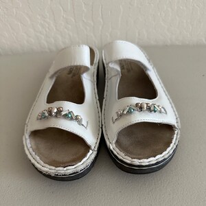 May include: A pair of white leather sandals with a jeweled embellishment on the toe strap. The sandals have a closed toe and an open heel.