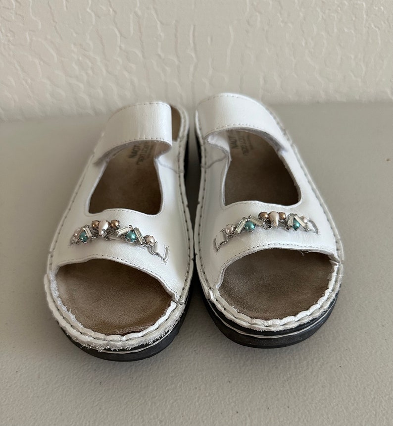 May include: A pair of white leather sandals with a jeweled embellishment on the toe strap. The sandals have a closed toe and an open heel.