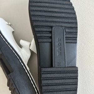 May include: White leather sandals with black soles and a black label that says "NAOE" and "38".