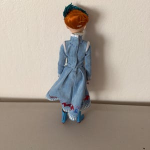 May include: A doll with red hair and a blue dress. The doll's dress has white trim and red and white accents. The doll is wearing blue boots and a green headpiece. The doll is standing on a light gray surface.