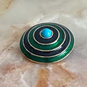 May include: A round, enameled brooch with concentric circles of black, green, and gold. The center features a small, round, turquoise-colored stone. The brooch has a shiny, polished finish and is set against a neutral background.