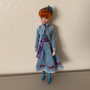 May include: A doll of a character with red hair, wearing a blue dress and jacket. The dress has a pattern of red and white designs. The doll has blue boots and a decorative headpiece. The doll is standing in front of a white wall.
