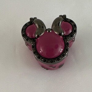 May include: A decorative ring in the shape of a mouse head. The ring features a dark gray metal frame with a deep pink center and accents. The frame is encrusted with small black stones.