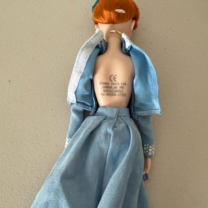 May include: A doll with auburn hair and a blue dress. The doll has a blue jacket with white trim and a blue skirt. The back of the doll has the text "Disney Store Ltd. LONDON, W6 9PE".