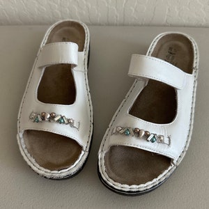 May include: A pair of white leather sandals with a decorative beaded strap. The sandals have a brown leather insole and a white stitched edge.