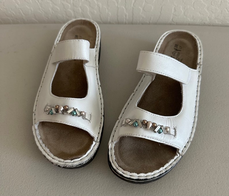 May include: A pair of white leather sandals with a decorative beaded strap. The sandals have a brown leather insole and a white stitched edge.
