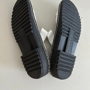 May include: A pair of white leather sandals with black soles and a black strap across the top. The sandals have a size 38 label on the sole.