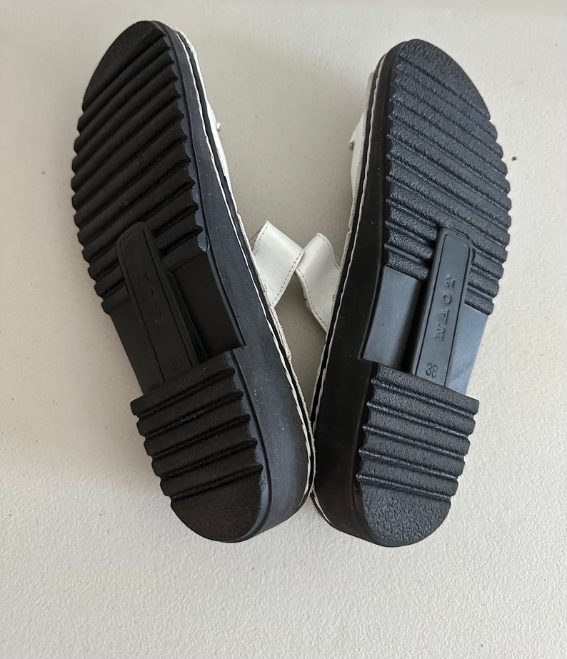 May include: A pair of white leather sandals with black soles and a black strap across the top. The sandals have a size 38 label on the sole.