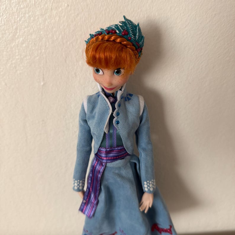 May include: A doll of a character with red hair, wearing a blue dress and jacket. The doll has a teal headpiece with red berries. The dress has a purple sash and the jacket has white trim. The doll is a collectible item.