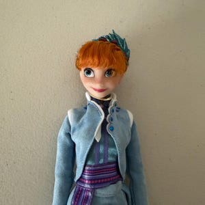 May include: A doll of a character with orange hair, blue eyes, and freckles. The doll wears a light blue jacket with white trim, a teal dress, and a purple and blue patterned sash. A green and red decorative piece adorns the doll's hair.