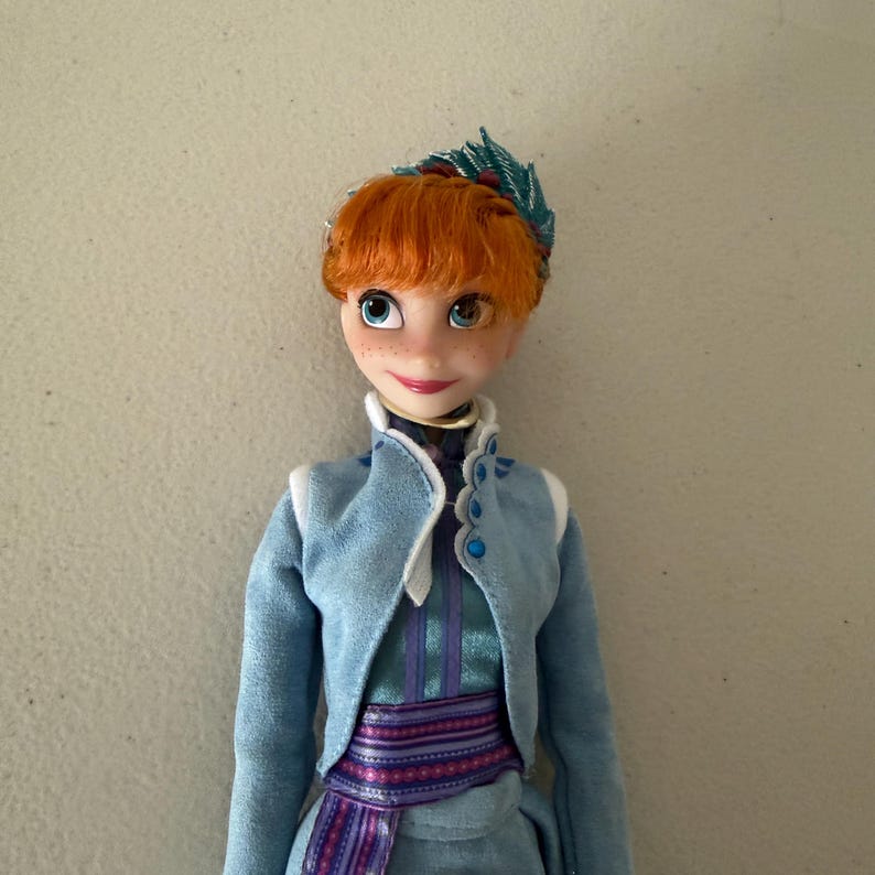 May include: A doll of a character with orange hair, blue eyes, and freckles. The doll wears a light blue jacket with white trim, a teal dress, and a purple and blue patterned sash. A green and red decorative piece adorns the doll's hair.