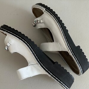 May include: A pair of white leather sandals with black soles and jeweled embellishments. The sandals have a strap across the toe and a strap around the ankle.