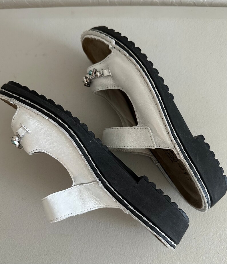 May include: A pair of white leather sandals with black soles and jeweled embellishments. The sandals have a strap across the toe and a strap around the ankle.