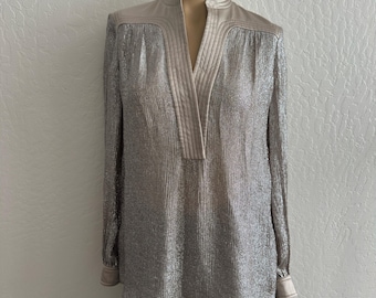 May include: A metallic silver long-sleeve blouse with a mandarin collar and a V-neckline. The blouse has a textured, shimmering appearance and is paired with black pants. The collar and neckline are a contrasting beige color.