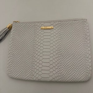 Peut inclure: Pochette en cuir blanc effet peau de serpent avec une fermeture éclair et un pompon dorés. La pochette porte un logo doré "Gigi New York".
