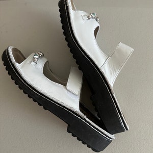 May include: A pair of white leather sandals with black soles and decorative embellishments. The sandals have two straps and an open toe.