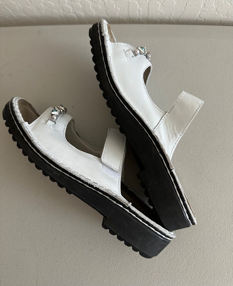 May include: A pair of white leather sandals with black soles and decorative embellishments. The sandals have two straps and an open toe.