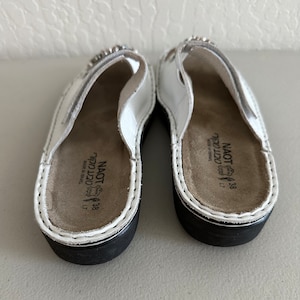 May include: A pair of white leather sandals with a black sole. The sandals feature a small rhinestone embellishment on the strap. The sandals are labelled "Naot" and "38" on the insole.