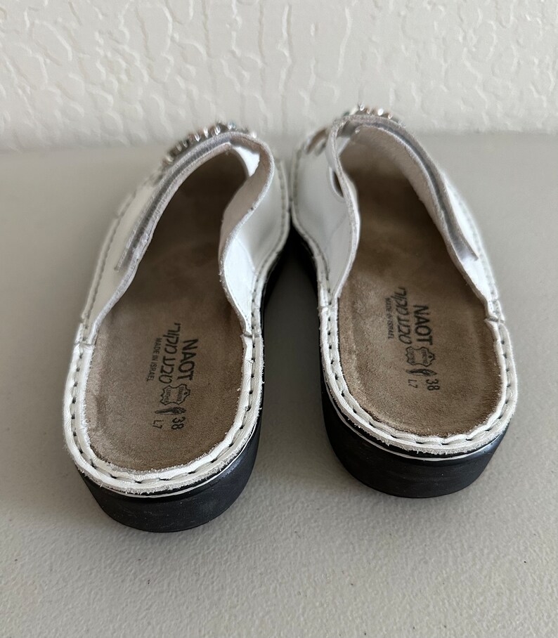 May include: A pair of white leather sandals with a black sole. The sandals feature a small rhinestone embellishment on the strap. The sandals are labelled "Naot" and "38" on the insole.