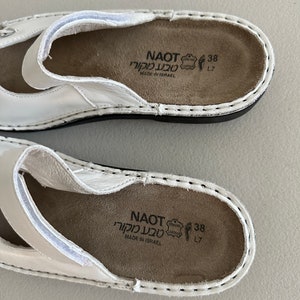 May include: Close-up of the inside of a white leather sandal with a brown insole. The insole has the text "NAOT טבע מקורי MADE IN ISRAEL" and the size "38 L7" printed on it.