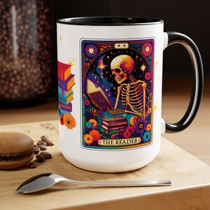 Puede incluir: Taza de cerámica blanca con borde y asa negros. La taza presenta una ilustración colorida de un esqueleto leyendo un libro con el texto "The Reader" en la parte inferior. La ilustración está rodeada de flores y estrellas.