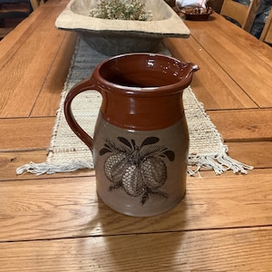 May include: A ceramic pitcher with a brown rim and handle, and a tan body. The body features a brown illustration of pinecones. The pitcher is sitting on a beige runner on a wooden table.